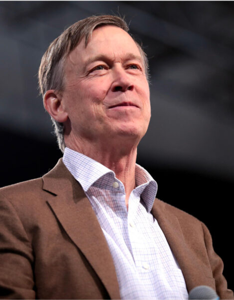 United States Senator for Colorado John Hickenlooper is looking up with a soft smile on his face. He is wearing a dark khaki suit and a white button down shirt.