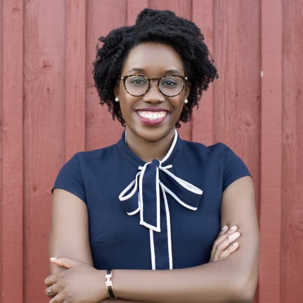 United States Representative Lauren Underwood is wearing glasses and smiling in front of a red wall. She is wearing a navy short sleeve blouse with a navy and white bow.