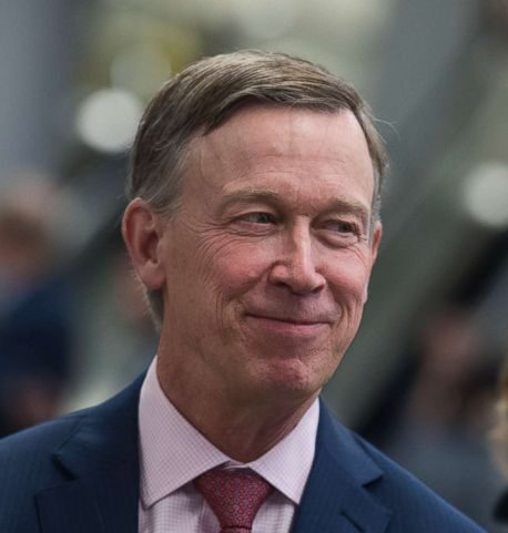 Headshot of United States Senator for Colorado John Hickenlooper smiling in a navy suit with a light pink dress shirt and red tie.