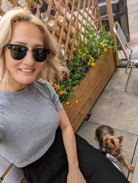 A blonde woman wearing black sunglasses and a gray shirt is taking a selfie in front of a flower bed.