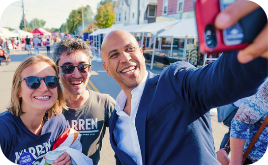 A smiling bald man in a navy blazer and a white button down shirt takes a selfie with a smiling man and woman wearing sunglasses and WARREN t-shirts.